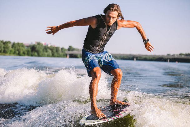Active wakesurfer jumping on wake board down the river waves. Surfer on wave. Male athlete training on wakesurf training. Active water sports in open air on board. A man catches a wave on surf. - Photo, Image