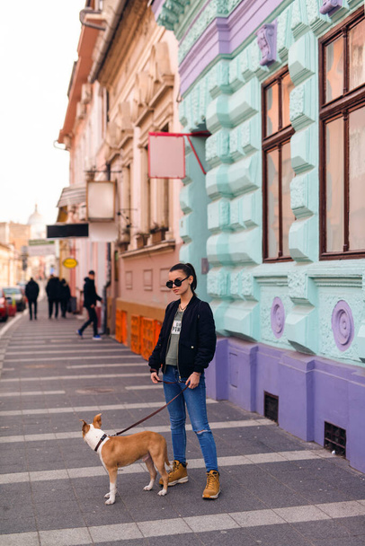 A beautiful caucasian girl with sunglasses stands next to her dog breed American Staffordshire Terrier in front of an old house on the street - 写真・画像