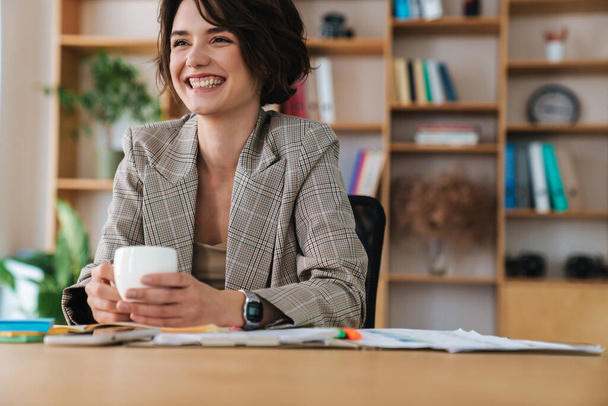 Bonita sorridente jovem empresária sentada na mesa do escritório, segurando xícara de café
 - Foto, Imagem