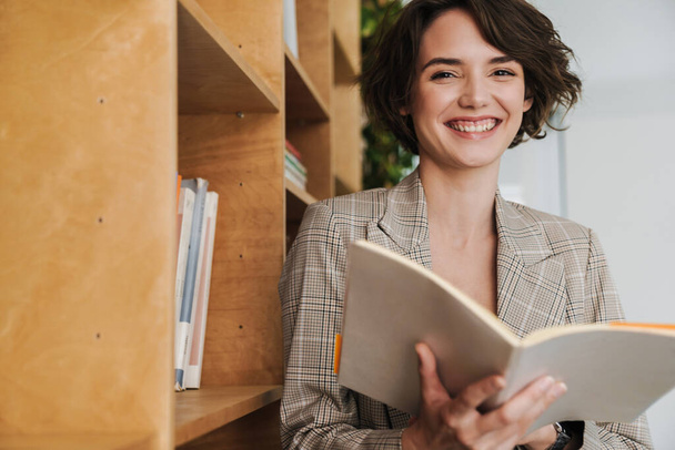 Smiling young woman entrepreneur reading book while standing in the office at the bookshelf - Photo, Image