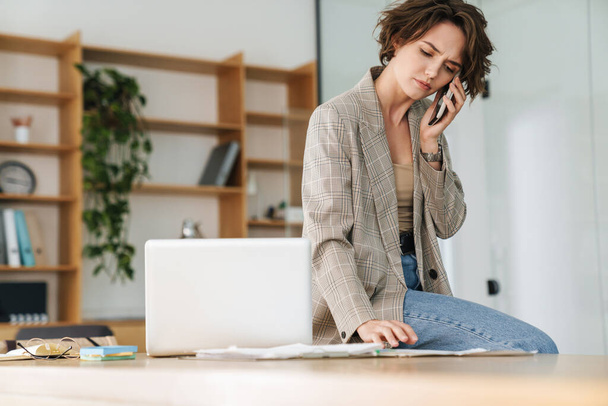 Attractive confident young businesswoman wearing jacket talking on mobile phone while sitting on a desk and using laptop computer in office - Foto, Bild