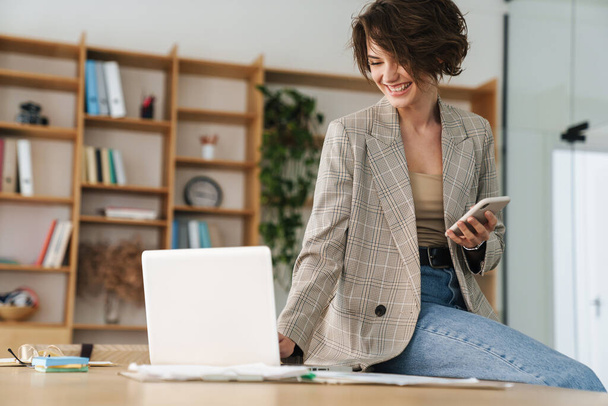 Attractive smiling young businesswoman wearing jacket holding mobile phone while sitting on a desk and using laptop computer in office - Фото, зображення
