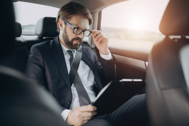 Mężczyzna czyta dokumenty siedząc na tylnym siedzeniu samochodu - Zdjęcie, obraz