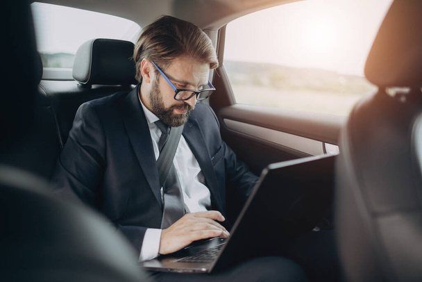 Hombre de negocios guapo sentado en el coche y trabajando en el ordenador portátil
 - Foto, imagen