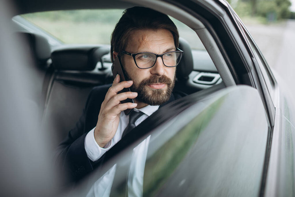Ο άνθρωπος κάθεται στο πίσω κάθισμα του αυτοκινήτου και μιλάει στο smartphone - Φωτογραφία, εικόνα