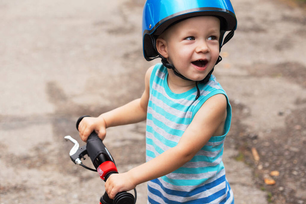 Onnellisesti smilling lapsi poika sininen paita ja kypärä seisoo tiellä tasapaino pyörä. - Valokuva, kuva