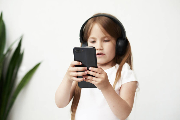 The little girl in headphones. The girl is listening music. The little girl is playing phone on the background. - Photo, image