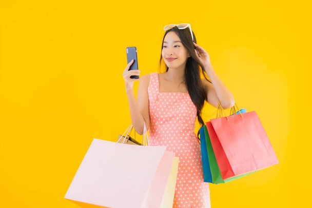 Retrato hermosa mujer asiática joven con colorido bolso de compras con teléfono móvil inteligente y tarjeta de crédito sobre fondo aislado amarillo
 - Foto, Imagen