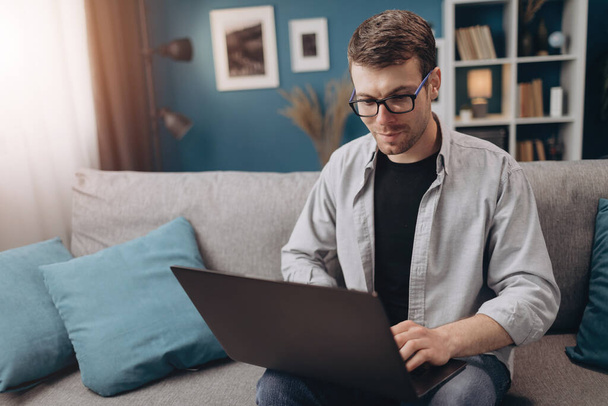 Щасливий хлопець використовує ноутбук, сидячи на сірому дивані
 - Фото, зображення