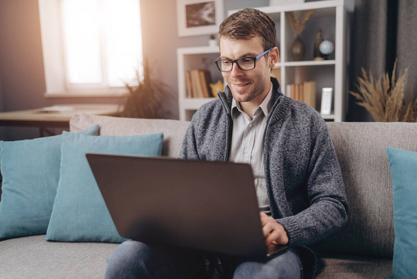 Usmívající se muž pracuje na notebooku, zatímco sedí na gauči - Fotografie, Obrázek