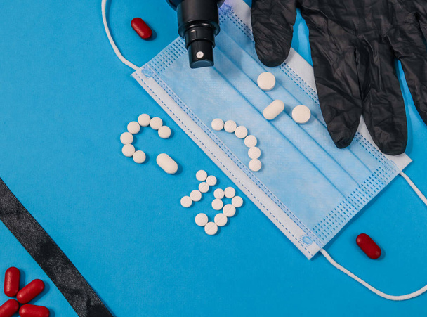 Masque médical, gants en noir, ruban noir et pilules en rouge et blanc sous la forme d'un émoticône triste et un symbole de coronavirus à droite sur un fond bleu et avec place pour le texte à gauche, cl - Photo, image
