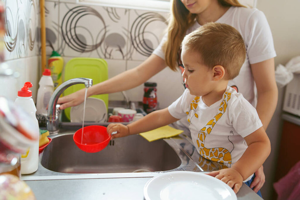 Вид сбоку на мать и сына на кухне дома - Молодая женщина с маленьким ребенком, моющая посуду - по пояс в день материнства и детства, растущего в понятии
 - Фото, изображение