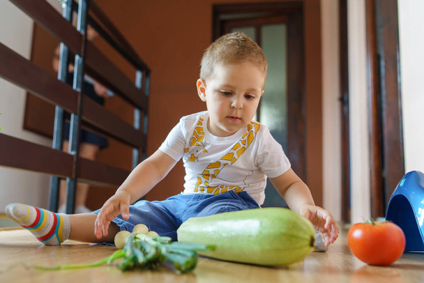 Vista frontale sul piccolo ragazzo caucasico seduto sul pavimento con verdure - Bambino che gioca con cipolla di pomodoro e zucchine a casa in sala - Infanzia e concetto di crescita - Foto, immagini