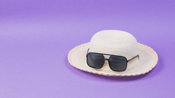Eflatun bir arka planda hasır bir şapka ve güneş gözlüğü.. - Fotoğraf, Görsel