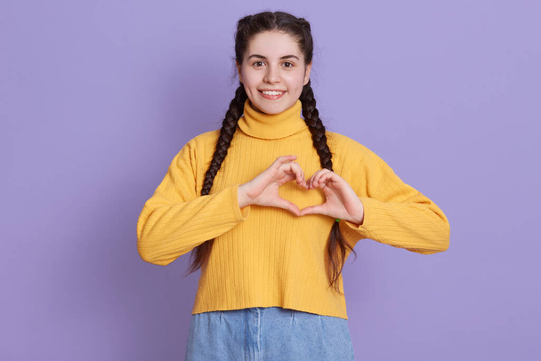 Attractive smiling girl with dark hair and pigtails, looking directly at camera with happy smile, making shape of heart with fingers, expressing love and gladness. - Foto, Imagen