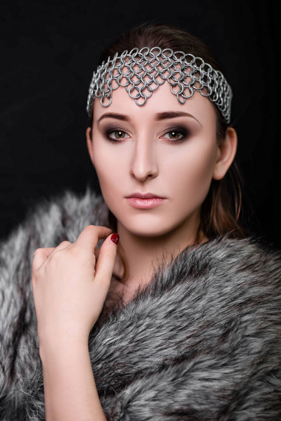 Beauty portrait of a woman with a chain mail bezel on her head and fur on her shoulders. Studio photo on a black background. Model with clear skin, retouch. - Photo, image