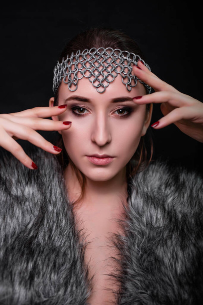 Beauty portrait of a woman with a chain mail bezel on her head and fur on her shoulders. Studio photo on a black background. Model with clear skin, retouch. - Foto, afbeelding