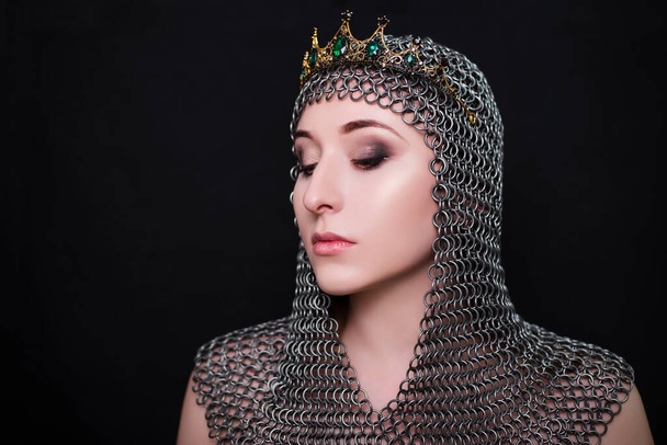 Beauty portrait of a woman in a chainmail hood with a crown on her head. Model with clear skin, retouching. Photo in studio on a black background. - Valokuva, kuva