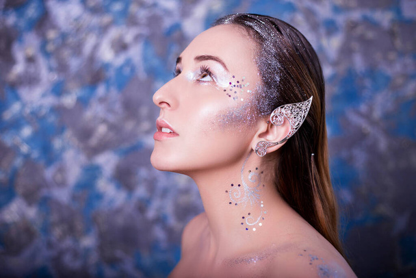Closeup beauty portrait of a woman with an earring on her ears. Studio photo on speckled, colorful background. Model with clean skin, retouching, makeup. The concept of elven beauty. - 写真・画像