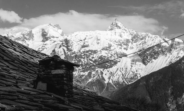 Een zwart-wit uitzicht op een schoorsteen boven de leistenen daken van een traditioneel Kinnauri huis tegen de achtergrond van de toppen van de Kinner Kailash range. - Foto, afbeelding