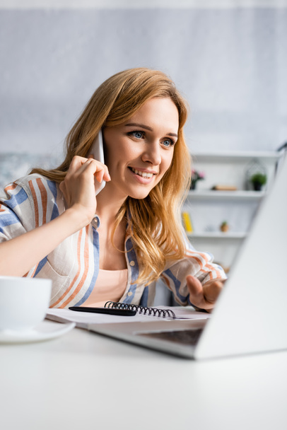 Concentration sélective de la femme souriante parlant sur smartphone et utilisant un ordinateur portable pendant l'entrevue d'emploi à la maison  - Photo, image