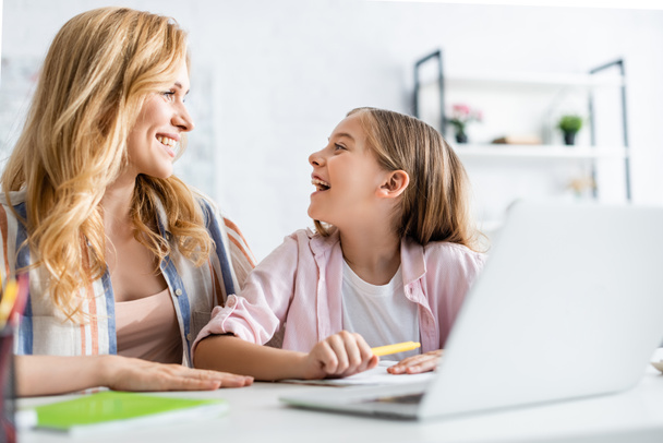 Селективное внимание матери, улыбающейся дочери возле канцелярских принадлежностей и ноутбука на столе дома
  - Фото, изображение