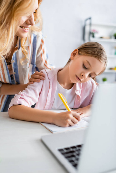Selective focus of smiling woman looking at kid writing on notebook near laptop  - Photo, Image