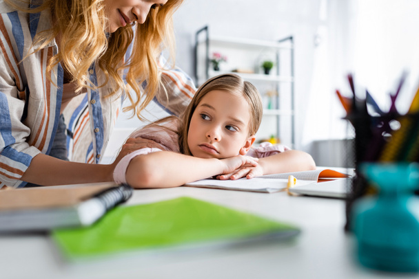 Selective focus of mother embracing sad daughter near stationery on table  - Foto, imagen