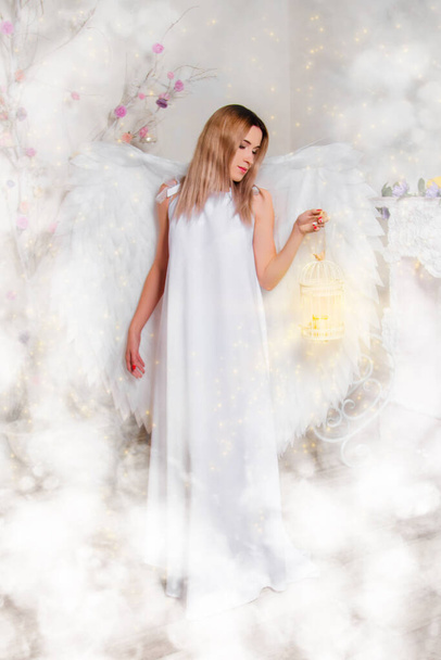 Una joven en una túnica blanca con grandes alas blancas detrás de su espalda está de pie en nubes blancas y sostiene una jaula de lámpara con luz mágica en sus manos
. - Foto, Imagen