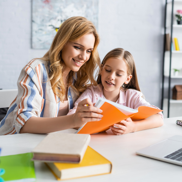 Selective focus of smiling mother and kid looking at notebook near laptop and books on table  - Photo, Image