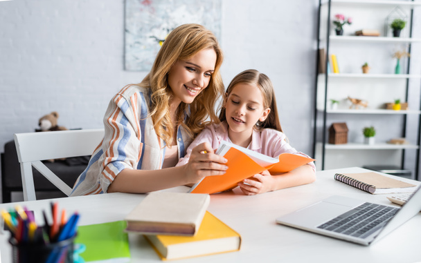 Selective focus of smiling woman holding notebook while helping daughter during online education at home   - Zdjęcie, obraz