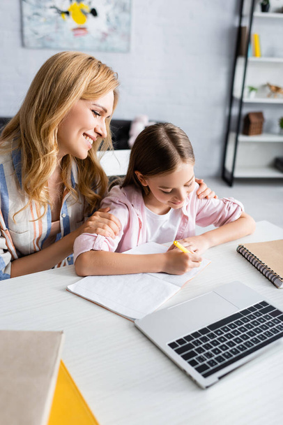 Selective focus of smiling woman embracing kid writing on notebook near laptop on table  - Foto, imagen
