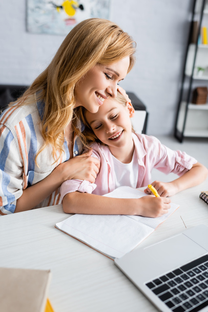 Foco seletivo da mulher abraçando a filha sorridente perto de artigos de papelaria e laptop na mesa em casa
  - Foto, Imagem