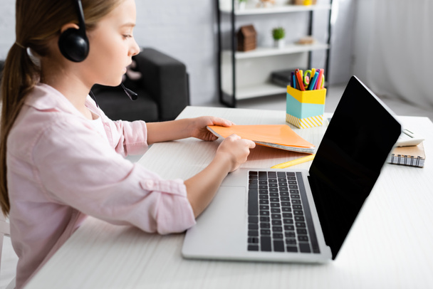 Widok z boku dziecka w słuchawkach biorąc notatnik w pobliżu laptopa na stole   - Zdjęcie, obraz