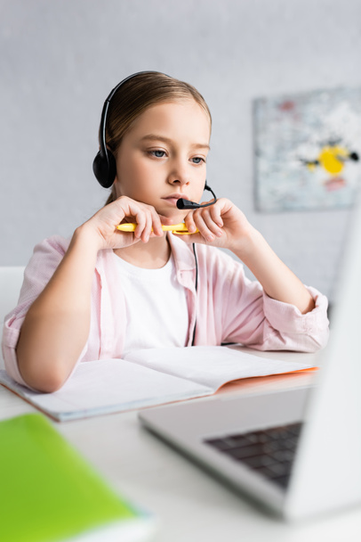 Selektywne skupienie uwagi dziecka w słuchawkach trzymających długopis i patrzących na laptopa  - Zdjęcie, obraz