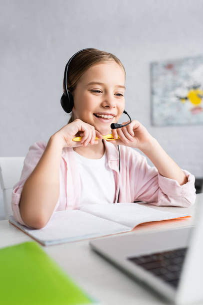 Selective focus of smiling child in headset holding pen and looking at laptop on table  - Photo, Image