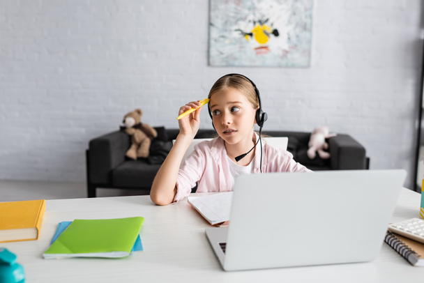 Selective focus of pensive child holding pen while using headset near laptop and stationery on table  - Fotoğraf, Görsel