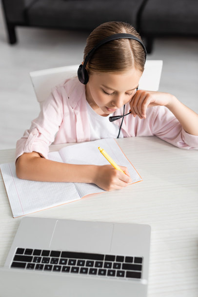Overhead view of kid in headset writing on notebook near laptop on table  - Fotó, kép