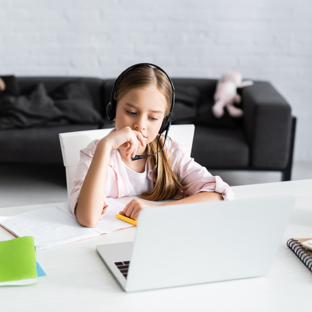Вибірковий фокус милої дитини в гарнітурі, що сидить поруч з ноутбуком і ноутбуком на столі
  - Фото, зображення