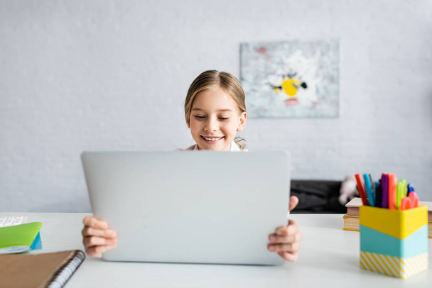 Concentration sélective d'un enfant souriant tenant un ordinateur portable près des livres sur la table  - Photo, image