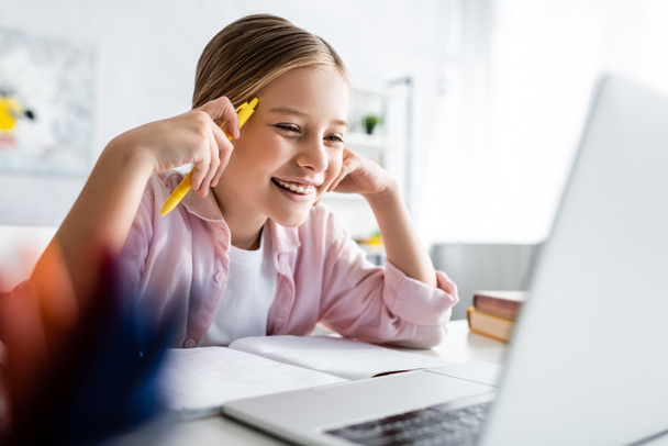 Селективный фокус положительного ребенка, держащего ручку рядом с ноутбуком и глядящего на ноутбук на столе
  - Фото, изображение