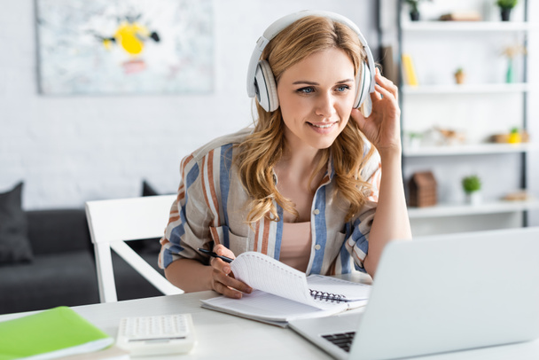 Selective focus of attractive woman in headphones looking at laptop while studying online - Фото, изображение