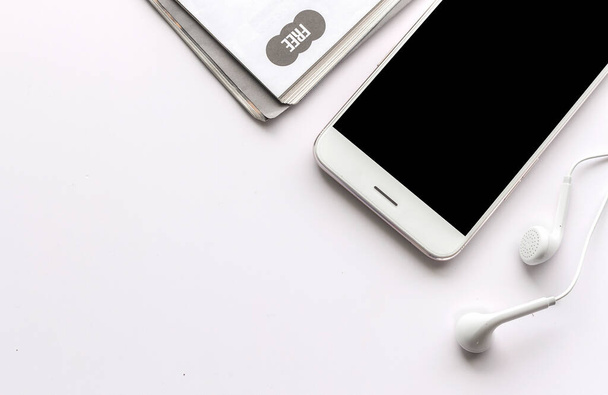 smartphone, sluchátka a časopis na bílém stole s více světla v pozadí - Fotografie, Obrázek