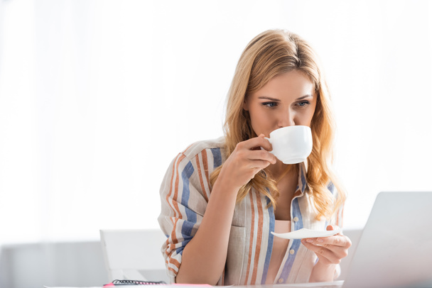 Selective focus of pretty woman drinking coffee while working with laptop - Photo, Image