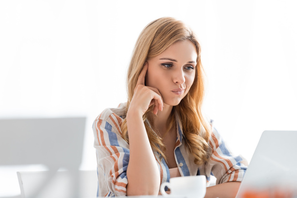 Selective focus of pensive woman looking at laptop with cup of tea - Photo, image