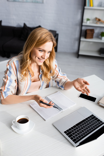 Selective focus of attractive woman smiling and gesturing near laptop and coffee - Foto, Imagen