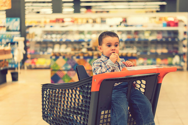 Roztomilý chlapec jí housku nebo chléb pita v supermarketu na vozíku. Hladové dítě v obchodě. tónovaný. - Fotografie, Obrázek
