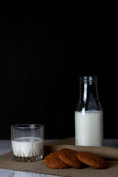 cookies, a bottle and a glass of milk on canvas napkin on white wooden background against black background. Vertical image - Fotoğraf, Görsel