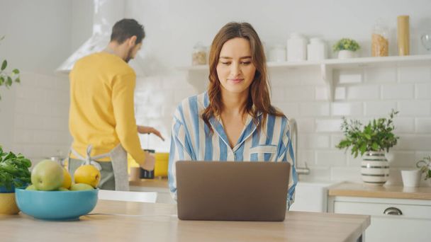 Young Beautiful Woman Using Laptop Computer While Wearing Blue Pyjamas. Brunette Female Sitting in a Modern Sunny Kitchen Room. Freelancer Working from Home. - Foto, immagini