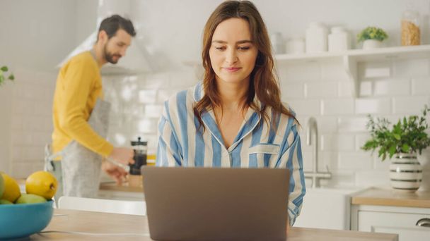 Young Beautiful Woman Using Laptop Computer While Wearing Blue Pyjamas. Brunette Female Sitting in a Modern Sunny Kitchen Room. Freelancer Working from Home. - Photo, Image